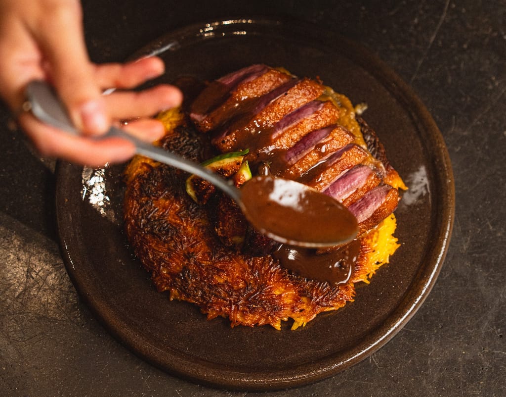 Viipaloitu ankka-annos rapean perunapohjan päällä, jonka päälle lusikoidaan tummaa kastiketta; annos kuvattu lähietäisyydeltä ravintolassa.