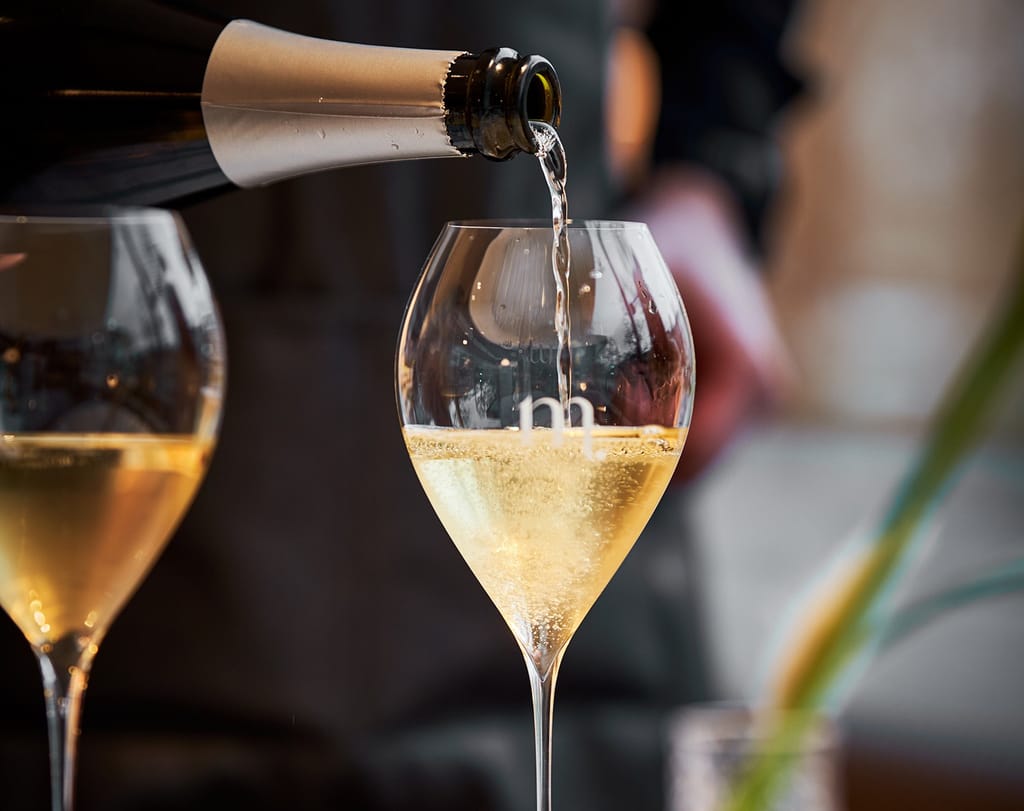 Kuohuviiniä kaadetaan pullosta korkeaan lasiin ravintolaympäristössä, taustalla toinen täytetty kuohuviinilasi.