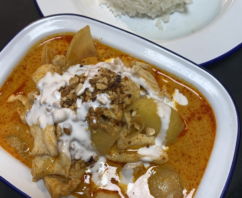 Kulhollinen kana-currya kookoskastikkeessa, päällä pähkinämurua, taustalla annos riisiä.