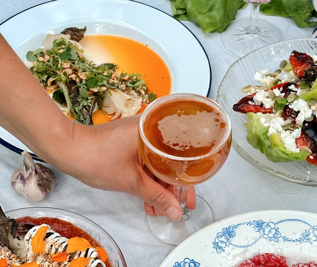 Käsi pitelee lasissa vaaleaa juomaa, pöydällä useita ravintola-annoksia valkoisilla lautasilla.