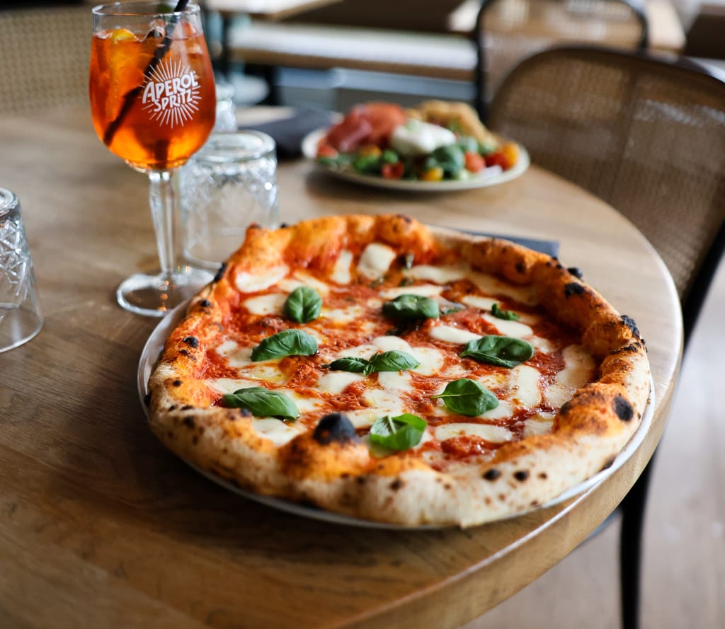 Puurakenteisella ravintolapöydällä on kiviuunissa paistettu margherita-pizza, jossa on rapea, hieman hiiltynyt reuna, tomaattikastiketta, sulanutta mozzarellaa ja tuoreita basilikanlehtiä. Pizzan vieressä on jalallinen lasi oranssia Aperol Spritz -juomaa jääpaloilla.