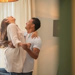 Mies nostaa naisen ilmaan ja pyörittää tätä iloisesti kotona. Nainen nauraa ääneen ja nojaa miehen olkapäähän, kun valo tulvii sisään verhojen läpi. Kuvassa näkyy spontaani onnellisuus, rakkaus ja läheinen yhteys kahden ihmisen välillä.