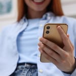 Sinipaitainen nainen hymyilee sille, mitä näkee puhelimensa ruudulta. Hänestä ei näy kuin leuka ja hampaat.