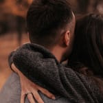 Pariskunta halaamassa ulkona syksyisessä puistossa, kuvattuna takaapäin; toisen käsi lepää kumppanin niskalla ja sormessa näkyy sormus.