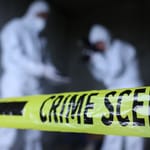 Kaksi valkoiseen suojapukuun pukeutunutta tutkijaa seisoo keltaisten poliisinauhojen takana mustaa taustaa vasten.