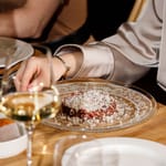 Ravintolapöydässä tarjottu annos ja viinilasit, kaksi ruokailijaa pöydän ääressä.