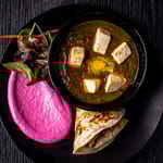 Ylhäältä kuvattu annos mustalla lautasella: vihreää currya tofukuutioilla, kirkkaanpinkkiä juurespyreetä, vihreää salaattia ja taiteltua naan-leipää tummalla taustalla.