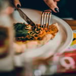 Lähikuva henkilöstä leikkaamassa pizzapalaan veitsellä ja haarukalla ravintolapöydässä; etualalla juomalasi ja taustalla näkyy Syö!-viikot-kampanjan materiaalia.