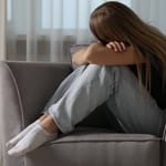 Nainen istuu sohvalla polvet rintaa vasten ja nojaa päätään käsiinsä, ilme kertoo ahdistuksesta.