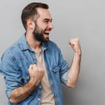 Iloinen parrakas mies nostaa nyrkit ilmaan riemuiten harmaata taustaa vasten.