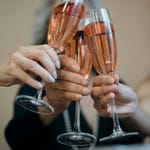 Kolme ihmistä kilistää vaaleanpunaista kuohuviiniä sisältäviä samppanjalaseja lähekkäin; kuvassa näkyy käsien lähikuva ja taustalla hymyilevät kasvot epätarkkoina.