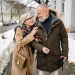 Kaksi aikuista kävelee talvisella jalkakäytävällä käsikynkässä, molemmilla on ulkovaatteet päällä ja lunta on maassa