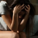 Nainen istuu etukumarassa ja pitelee päätään käsissään, ilme surullinen ja ahdistunut.