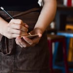 Tarjoilija kirjoittaa tilausta muistilapulle kynällä, yllään ruskea esiliina ja taustalla ravintolasali epätarkkana.