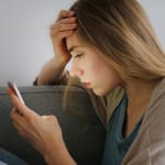 Huolestunut nainen istuu sohvalla ja katsoo puhelintaan käsi otsalla, lähikuva sivuprofiilista vaaleaa seinää vasten.