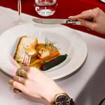 Henkilö leikkaa veitsellä ja haarukalla annosta, jossa on paistettua kalaa, kasviksia ja kastiketta valkoisella lautasella ravintolapöydässä.