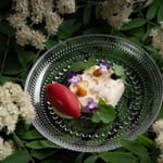 Taiteellisesti aseteltu jälkiruoka lasilautasella, ympärillä kukkia ja vihreitä lehtiä.