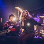 Nuori laulaja esiintyy hymyillen pienen bändin kanssa lavalla, taustalla rumpusetti ja kitaristeja.