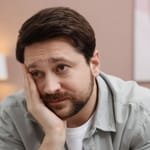 Mies nojaa käteensä ja katsoo hieman väsyneenä sivulle sisätiloissa.