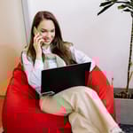 Nainen istuu säkkituolissa, puhuu puhelimessa ja käyttää kannettavaa tietokonetta.