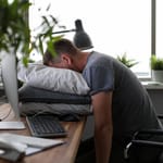 Uupunut mies nukkuu työpöydän ääressä tyynyjen päällä.