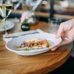 Tarjoilija asettaa ravintolassa pöytään annoksen, jossa on koristeellinen esillepano ja viinilasit taustalla.