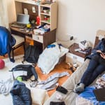 Sotkuinen nuoren huone, lattialla vaatteita ja tavaroita, henkilö makaa sängyllä puhelin kädessä.