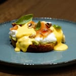 Annos lautasella: paahdetun leivän päällä avokadoa, uppomuna, savulohta ja hollandaise-kastiketta, koristeltu yrteillä ja pähkinöillä.