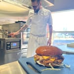 Etualalla kanaburgeri, taustalla kokki valmistaa ruokaa.