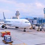 Suuri matkustajalentokone portilla lentokenttäterminaalissa, taustalla lennonjohtotornit ja kuljetuskontteja asfaltilla.