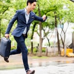 Kiireinen liikemies juoksee sateisella kadulla katsoen kelloa, myöhässä tapaamisesta