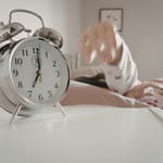 Herätyskello yöpöydällä, käsi kurottaa sitä kohti sängystä.
