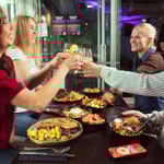 Ravintolapöytä täynnä ruokia. Ihmiset kilistävät laseja.