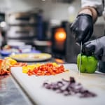 Kokki pilkkoo värikkäitä paprikoita ammattikeittiössä mustat hanskat kädessä