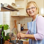 Hymyilevä keski-ikäinen nainen siniraitaisessa esiliinassa sekoittaa puulusikalla kattilassa kotikeittiössä