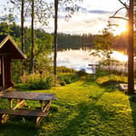 Suomalainen kesämökki järven rannalla iltaruskon valossa, edessä nurmikko ja piknikpöytä, taustalla vene ja koivut.