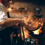Ammattikokki paistaa wokkipannulla avotulen äärellä savuisessa ravintolakeittiössä