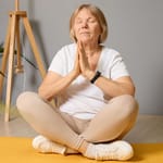 Iäkkäämpi nainen istuu ristijaloin joogamatolla silmät suljettuina, kädet rinnalla meditaatioasennossa