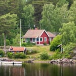 Punainen puinen kesämökki tiilikattoineen sijaitsee vehreässä metsäisessä rinteessä kallioisella järven- tai merenrannalla.