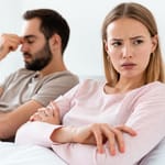 Pariskunta sängyssä selin toisiinsa riidan jälkeen – nainen risti käsillä, mies hieroo otsaansa stressaantuneena