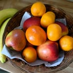 Hedelmäkorissa keittiön pöydällä on omenoita, mandariineja ja mangoja, ja korin vieressä lepää kaksi keltaista banaania.
