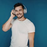 Hymyilevä mies kuuntelee musiikkia suurilla kuulokkeilla sinistä taustaa vasten, mikä kuvastaa äänen havainnointia ja äänikokemusta.