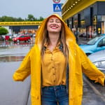 Keltainen sadetakki päällään oleva nainen seisoo hymyillen silmät suljettuina kesäisessä vesisateessa parkkipaikalla autojen keskellä.