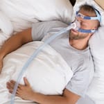 Mies nukkuu selällään CPAP-laitteen maski nenällään.