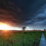 Dramaattiset ukkospilvet ja kirkas auringonpaiste suomalaisella niityllä, lankkupolku johtaa horisonttiin.
