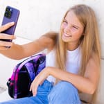 Hymyilevä pitkähiuksinen nuori tyttö ottaa selfietä puhelimellaan nojaten seinään koulureppu vieressään.