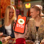 Henkilöitä syömässä ravintolassa. Käsi pitelee puhelinta, jossa näytöllä SYÖ!-viikkojen logo.