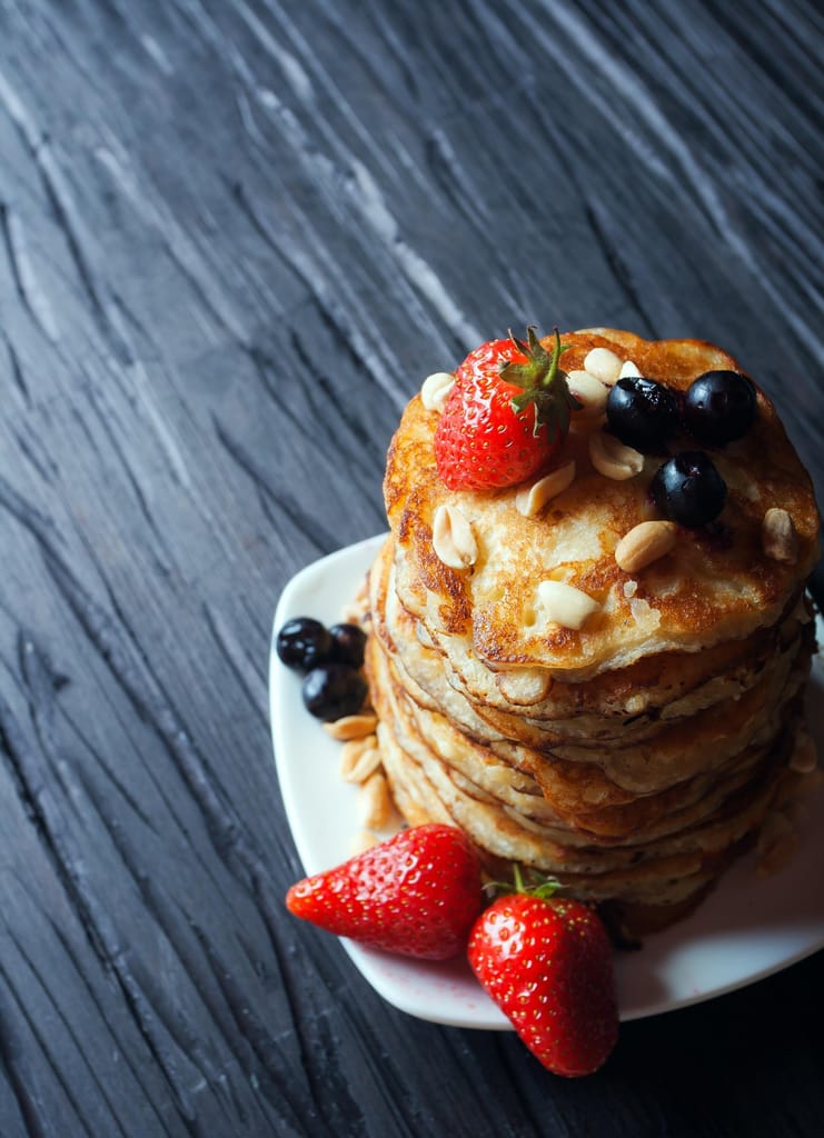 Kuvassa pino pannukakkuja harmaalle pöydälle asetetulla lautasella. Pannukakkujen päällä ja lautasella on marjoja.