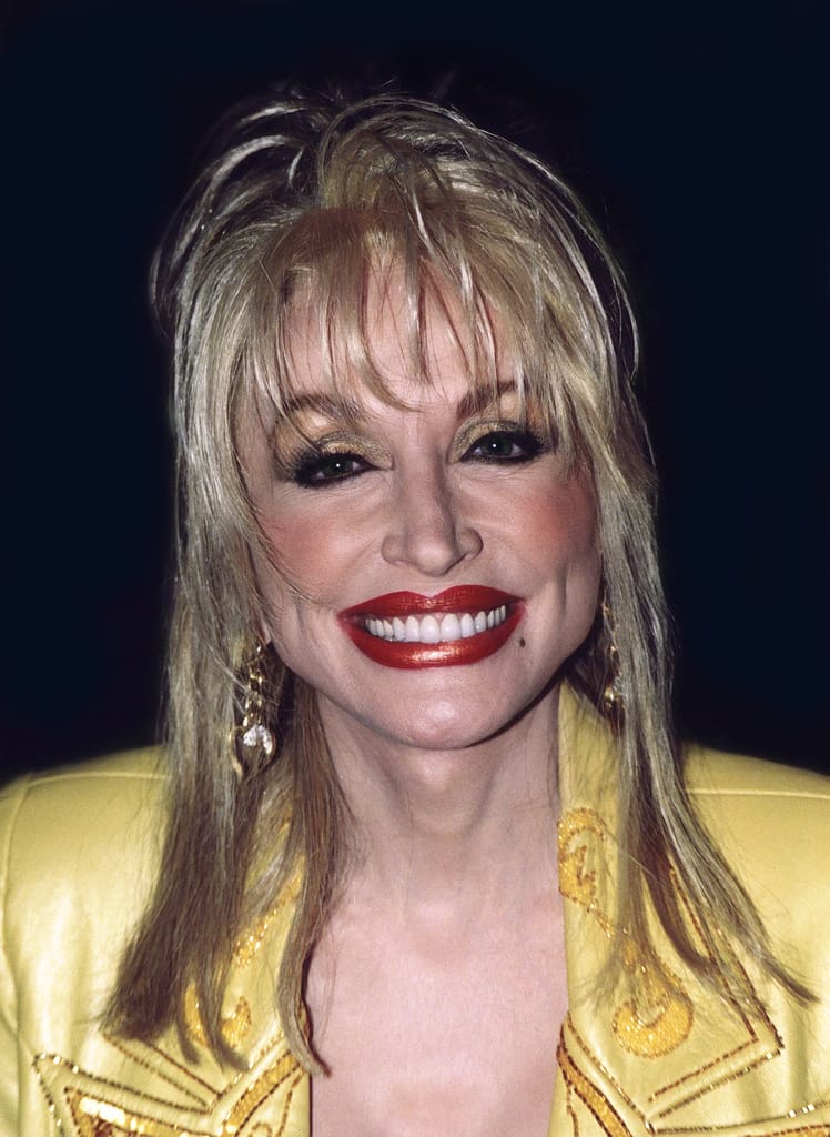 Dolly Parton hymyilee kameralle keltaisessa asussa, punaisessa huulipunassa ja kultaisissa korvakoruissa.