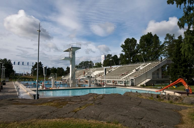 Helsingin Uimastadionin hyppytorni ja lastenallas vesiliukumäkineen, etualalla kallio ja taustalla katsomo ja siniset liput.